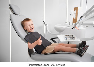 Horizontal side view snapshot of a little smiling boy wearing black bib, sitting in dentist chair, thumb up. Well equipped dentist office. Children's dentistry and medicine concept - Powered by Shutterstock