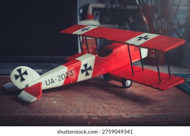 Horizontal side-angle view of a handcrafted red and white wooden triplane model with a propeller, resting on a wooden workbench in a workshop.
