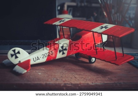 Horizontal side-angle view of a handcrafted red and white wooden triplane model with a propeller, resting on a wooden workbench in a workshop.
