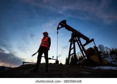 Horizontal snapshot of a silhouette of an oilman in orange vest and helmet, standing with his back to the oil pump jack in oilfield holding a pipe wrench, looking away, evening sky on background - Shutterstock ID 1803586168