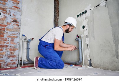 Horizontal Snapshot Of Young Plumber Working With Grey Sewer Pipes, Fixing Them To Wall With A Help Of Screwdriver. Side View Of Plumber Standing On Knees Wearing Blue Uniform And White Helmet