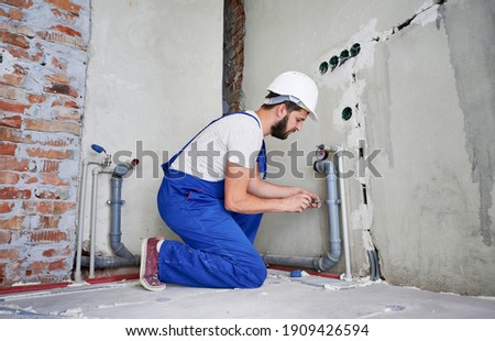 Horizontal snapshot of young plumber working with grey sewer pipes, fixing them to wall with a help of screwdriver. Side view of plumber standing on knees wearing blue uniform and white helmet