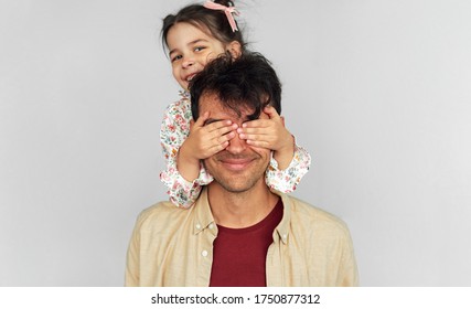 Horizontal Studio Image Of Cute Daughter Covers The Eyes With Hands Of Her Father For A Surprise On Father's Day. Handsome Dad Playing With His Little Girl Kid Peekaboo Game, Shares Love Together.