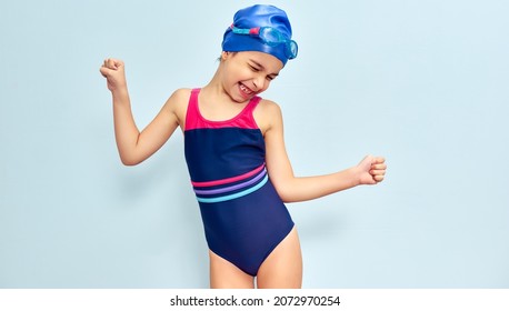 Horizontal Studio Image Of A Happy Little Girl In A Swimsuit, Swim Goggles And Swim Cap Making Gesture During She Wins The Swimming Contest, Isolated Over Blue Studio Background.