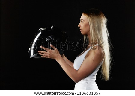 Horizontal studio image of young blonde woman wearing white tank top holding black motor helmet. Attractive sporty girl posing isolated with protective equipment. Extreme sports and transportation