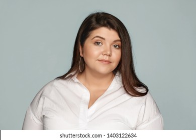 Horizontal Studio Portrait Of Adorable Beautiful Young Overweight European Woman With Loose Black Hair And Chubby Cheeks Wearing Round Earrings And Xxl White Shirt Posing At Blank Wall, Smiling