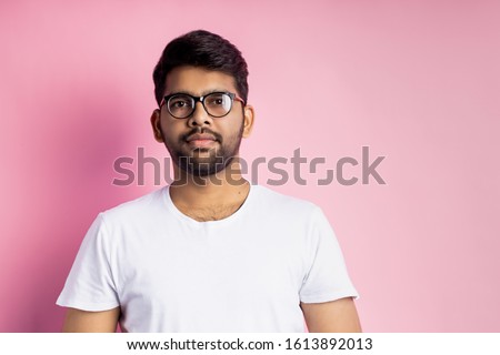 Horizontal studio portrait of young handsome friendly indian unshaven man, wearing glasses, white t shirt, looking at camera with calm expression, isolated over pink background.