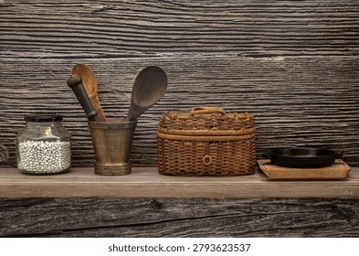 A horizontal studio shot of a rustic kitchen still life arrangement on a weathered wooden shelf against a textured dark wood background. From left to right: a glass jar filled with white cannellini beans, an antique brass mortar and pestle holding two carved wooden spoons, a handwoven wicker basket with a lid, and a small seasoned cast iron skillet on a wooden trivet.