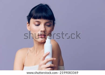 Horizontal studio shot.A beautiful joyful woman in a white T-shirt with clean beautiful well-groomed skin, black wavy short hair gathered in a ponytail stands on a lilac background with a bottle