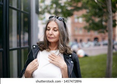 Horizontal Summer Image Of Attractive Serious Middle Aged Retired Woman Posing Outdoors, Standing At Modern Building Dressed In Stylish Black Leather Jacket, Going To Get Inside Cafe Or Shop