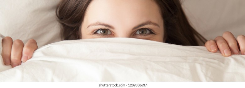 Horizontal top close up shy playful woman looking smiling eyes at camera hiding under white blanket lying on pillow in bed having fun concept, banner for website header design with copy space for text - Shutterstock ID 1289838637