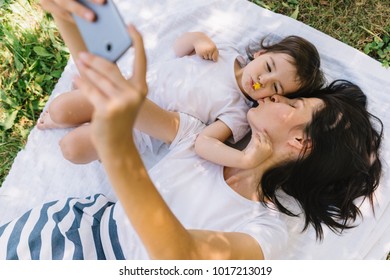 Horizontal top view of beautiful young mother blowing kiss, taking self portrait with beautiful little girl daughter lying on white plaid on grass. Young family taking selfie outdoor. Motherhood - Powered by Shutterstock