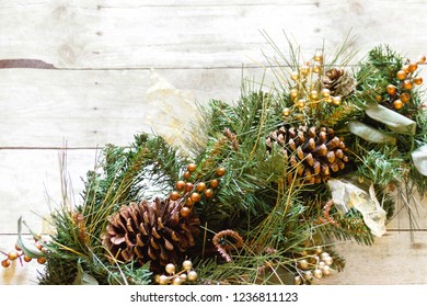 Horizontal top-down view of an offset arrangement of holiday greenery with fir branches, pinecones, gold covered cranberries, leaves, and sheer gold ribbon on top of a gray wooden backdrop - Powered by Shutterstock