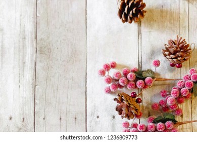 Horizontal top-down view of a small group of red frosted cranberries and pinecone ornaments scattered on the right hand side of a gray wood plank backdrop