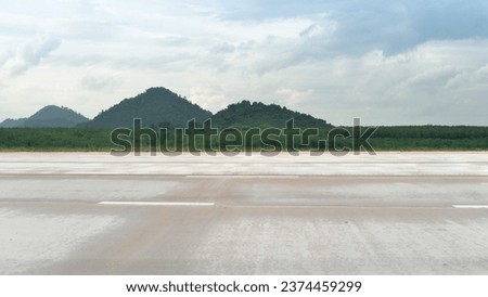 Horizontal view of empty concrete road with dust and grass. Background of green forest and mountain under tha sky with clouds.