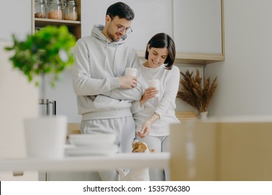 Horizontal view of happy young woman and man play with dog, drink takeaway coffee, stand against kitchen interior, unpack boxes with personal stuff, wear casual clothes, bought new apartment - Powered by Shutterstock