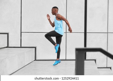 Horizontal view of male athlete dressed in activewear, has cardio running up stairs prepares for jogging on long distance makes steps or jumps high. African American man in t shirt, leggings, trainers