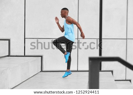 Horizontal view of male athlete dressed in activewear, has cardio running up stairs prepares for jogging on long distance makes steps or jumps high. African American man in t shirt, leggings, trainers