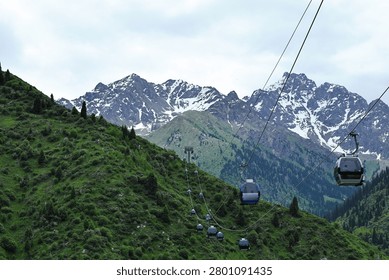 Horizontal view of multiple cable car cabins moving along lines over green summer slopes with majestic rocky snow-capped peaks in the background.