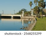 Horizontal View north from Sunset Park in Saint Petersburg, Florida with a leading line of seawall with stones, grass and palm trees on right. Central Avenue Bridge on left. Late afternoon sun reflect