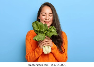 Horizontal View Of Pleased Korean Woman Holds Freshly Leafy Vegetable, Enjoys Eating Nutrient Food Containing Vitamins, Keeps Eyes Closed With Pleasure, Wears Orange Sweater, Isolated On Blue Wall