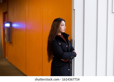 Horizontal View Of Pretty Brunette Woman With Waist Length Hair Wearing Black Coat Standing With Arms Crossed And Dejected Expression At The Entrance Of An Indoor Parking Building