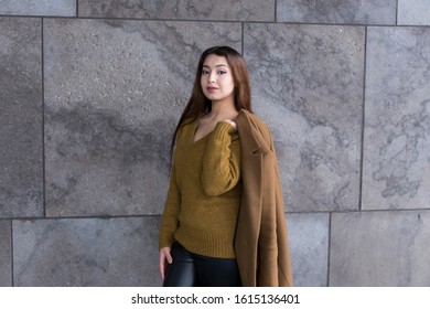 Horizontal View Of Pretty Brunette Woman With Waist Length Hair In Mustard Coloured Sweater Standing In Front Of Stone Wall Holding Her Light Brown Coat On Her Shoulder