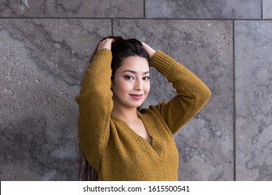 Horizontal View Of Pretty Smiling Brunette Woman In Mustard Coloured Sweater Standing In Front Of Stone Wall With Arms Raised Lifting Her Thick Waist Length Hair