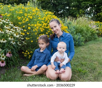Horizontal View Of Pretty Smiling Brunette Young Woman Kneeling In Grass Holding Serious Baby, With Older Sister Looking Forlorn Next To Her, Quebec City, Quebec, Canada