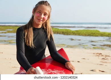 Horizontal View Of Professional Surfer Gets Ready For Surf, Waxes Board With Wax, Wants To Demonstrate Double Spinner, Has Sunblock Cream On Face, Wears Waterproof Bathingsuit, Models On Coast