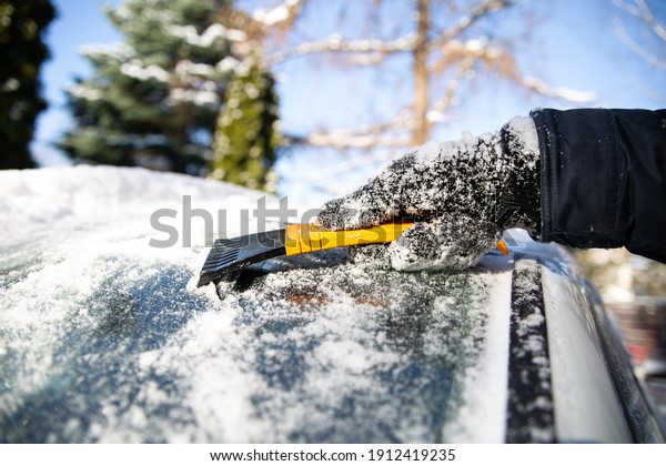 Horizontal view of\
snow clearing a car in winter. A hand in a glove cleaning a\
windshield of the car with a plastic scraper. Snow and ice removal\
from an auto, outdoor, on a sunny\
day.