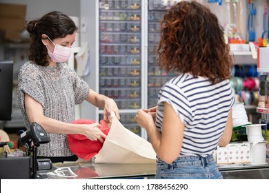 Horizontal View Of Woman Buying Clothes On A Retail Shop. Assistant At The Counter Wearing A Protective Face Mask To Avoid Covid 19 Contagious.