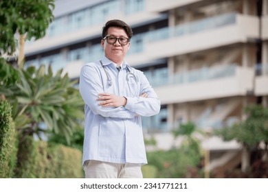 Horizontal waist-up portrait of a handsome male Arab Malay doctor, wearing white coat, standing with arms crossed outdoors on the background of clinic building. Close up, copy space - Shutterstock ID 2341771211
