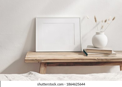 Horizontal white frame mockup on vintage wooden bench, table. Modern white ceramic vase with dry Lagurus ovatus grass and books. White wall background. Scandinavian interior. Selective focus.