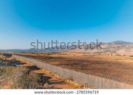 Horizontal wide angle shot of a secured border zone with a metal wire fence, military outposts, rural hillside villages, and terraced agricultural fields under a bright blue sky