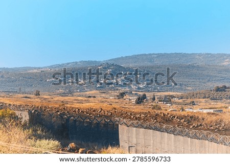 Horizontal wide angle shot of a secured border zone with a metal wire fence, military outposts, rural hillside villages, and terraced agricultural fields under a bright blue sky