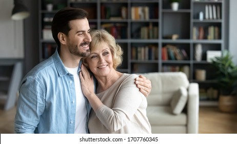 Horizontal wide image inside of cozy living room at home standing hugging middle-aged mother and grown up adult millennial son. Family bonds and values, love care and relatives closest people concept