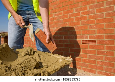 A horizontally cropped shot of a bricklayer in safety vest and denim applying mortar to a new brick using a trowel