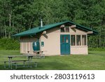 A horizontally-oriented photograph of a charming, rustic park shelter or public cabin building nestled in a grassy field. The building features light tan textured walls, a bright green metal roof with a small chimney pipe, a matching dark green wooden entrance door, and a large screened or windowed porch section. A distinctive green rain barrel sits beneath a downspout on the left side of the structure. In the foreground, wooden picnic tables with metal frames rest on the manicured lawn, while a dense forest of lush green deciduous trees forms a vibrant natural background. This high-quality image captures a peaceful summer day in a park or campground, making it ideal for travel brochures, outdoor recreation marketing, environmental design assets, or camping and tourism promotions.