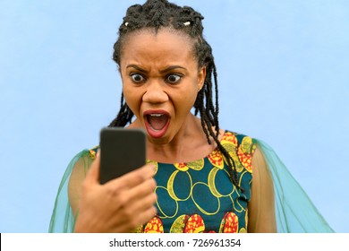 Horrified Upset Young African American Woman In A Stylish Dress Gawping Wide Eyed At Her Mobile Phone Isolated Over Blue