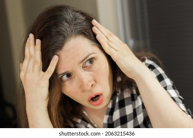 Horrified young woman looking in the bathroom mirror staring open mouthed at the first grey hair on her scalp, a first sign of ageing, or noticing that she is suffering from dandruff.