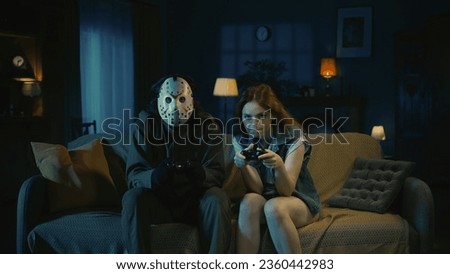 Horror maniac in white mask playing video games with a young girl, sitting on the sofa in the dark apartment room.