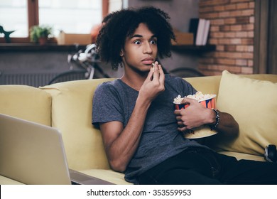 Horror Movie. Young African Man Watching TV And Looking Scared While Eating Popcorn Sitting On The Couch At Home