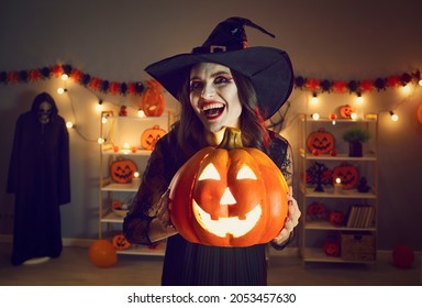 Horror Night. Halloween Portrait Of Spooky And Evil Witch With Glowing Jack-o-lantern In Hands. Young Woman In Witch's Hat With Crazy And Creepy Smile Looks Into Camera While Standing In Dark Room.