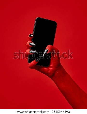 Horror red devil's hand with blank long nails holding smartphone with blank screen against red studio background. Concept of season sale, promotion, celebration, web. Negative space to insert text. Ad