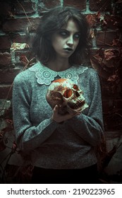 Horror Scene. A Sinister Ghost Girl With Faded Eyes Stands In A Gloomy Room Holding A Burning Human Skull In Her Hands. Halloween.