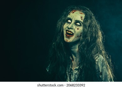 Horror, Thriller. Halloween. A Horrible Zombie Woman With Faded Eyes Bares Her Bloody Teeth. Devil Possessed Woman. Black Background With A Haze And Copy Space.