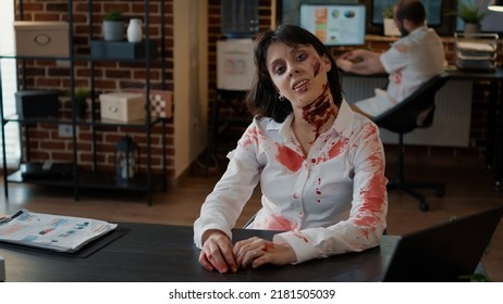 Horror Zombie Smirking Creepy In Office Workspace While Growling At Camera. Brain Dead Monster With Deep And Gorish Wounds Sitting At Table In Workspace With A Evil And Bizarre Smile.