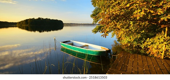 Horse Chestnut Tree by calm lake in full autumn colour at sunset and a rowing boat tied to a wooden pier