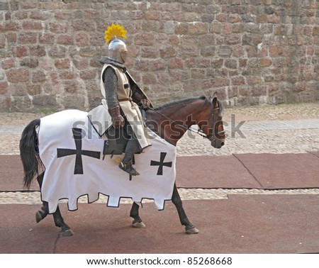The horse knight in an armor, going on a horse along a stone wall